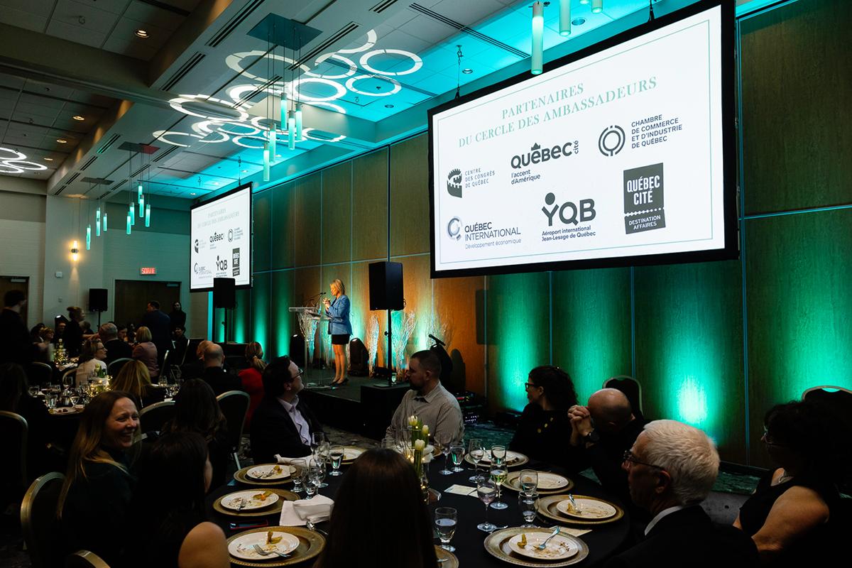 Audience seated at round tables during a conference banquet with a presentation on screen in Québec City.